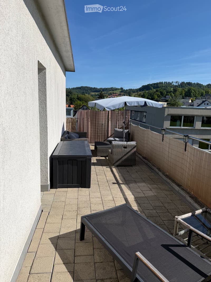 Balcony with tiled floor, wooden fence, sofa, table, chairs, sun lounger, and umbrella providing shade.