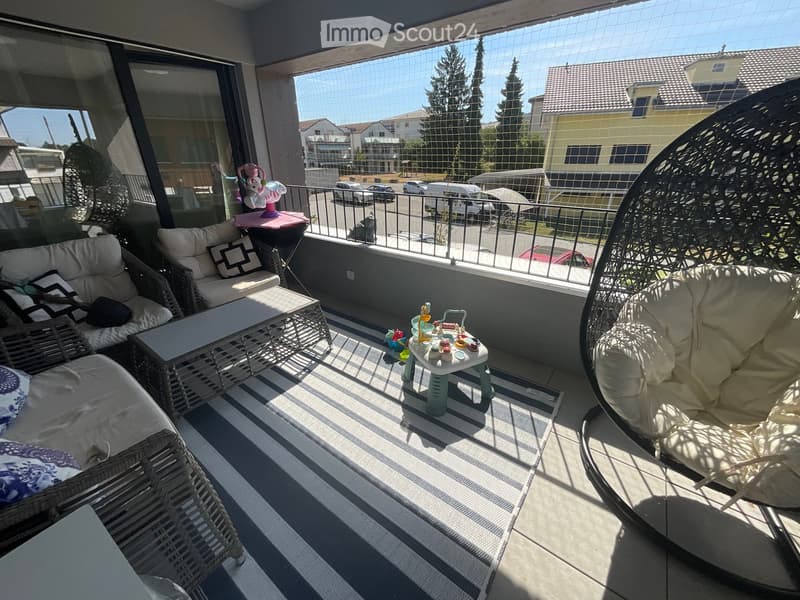 Balcony with wicker furniture, sofa, coffee table, and a small table with toys, overlooking a residential area with trees and parked cars.