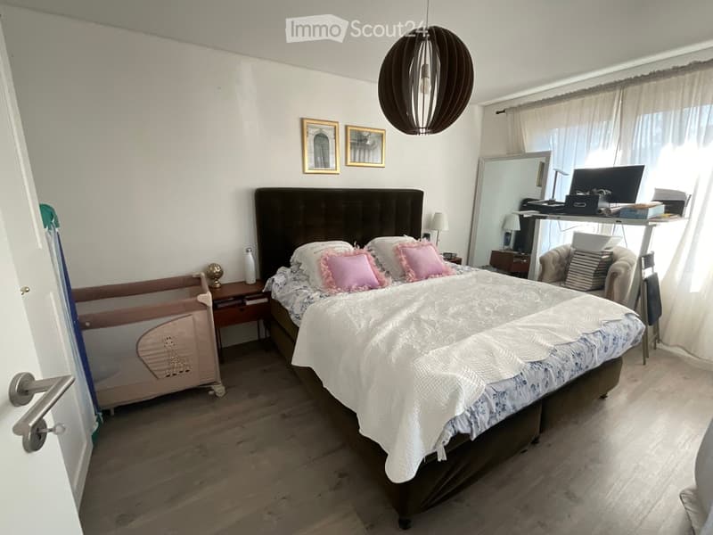 Cozy bedroom with a large bed, pink pillows, a wooden nightstand, a mirror, a chair, and a small table with a laptop and books.