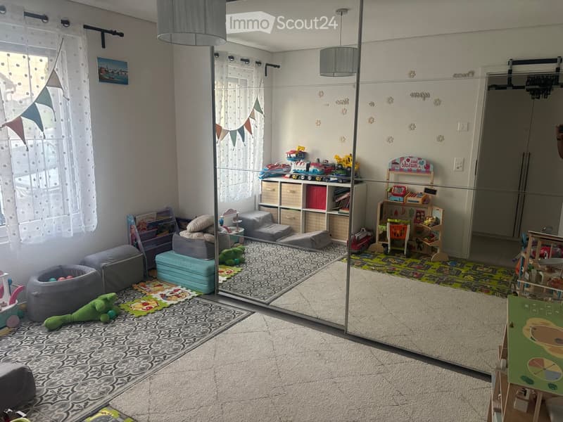 A well-lit, modern children's room with a large glass partition, colorful rugs, and various toys and furniture including a sofa, stools, and shelves.