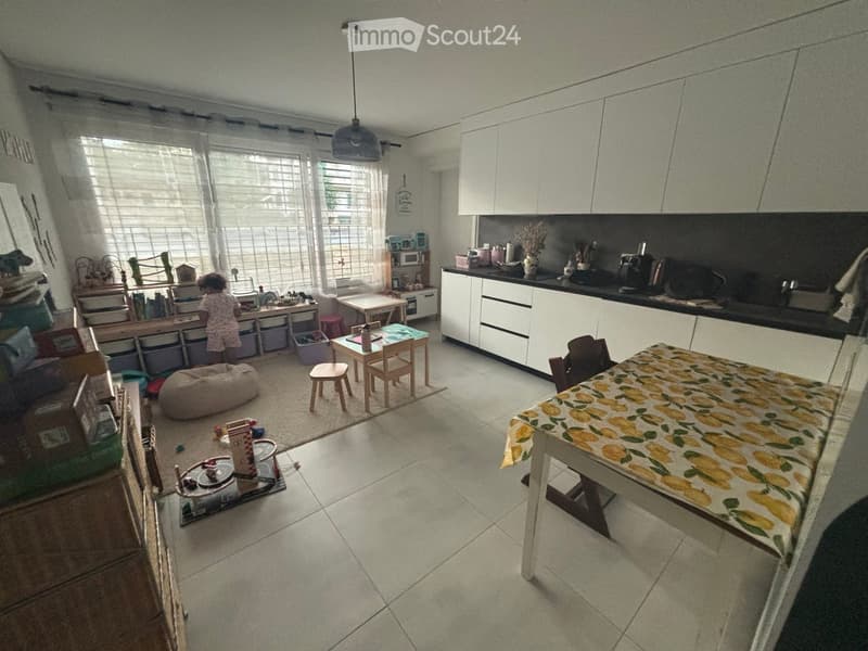 A combined living and kitchen area with a beige bean bag, small table and chairs, kitchen cabinets, and a dining table with chairs.