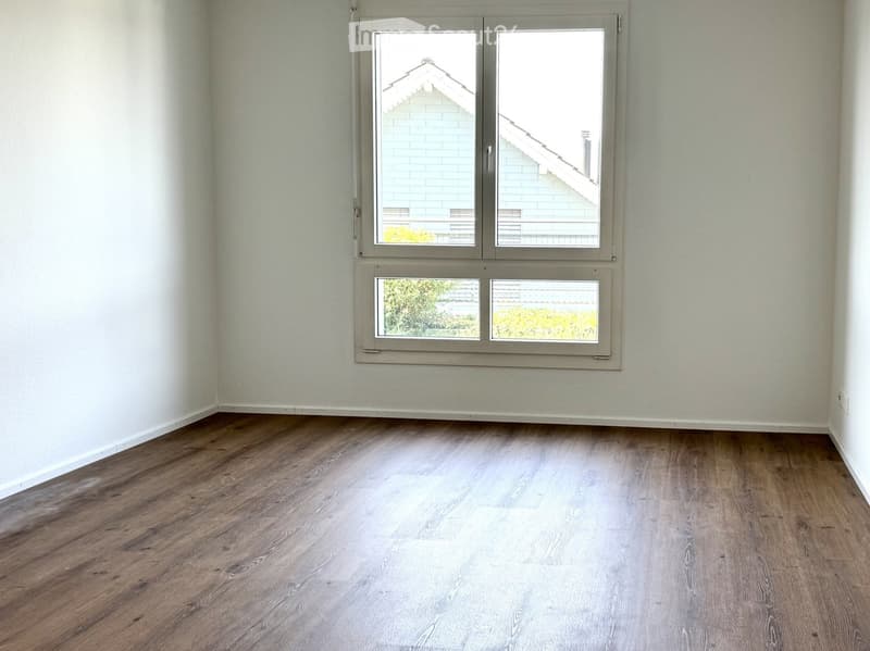 Empty room with light-colored wooden flooring and white walls, featuring two windows with grey frames and blinds.