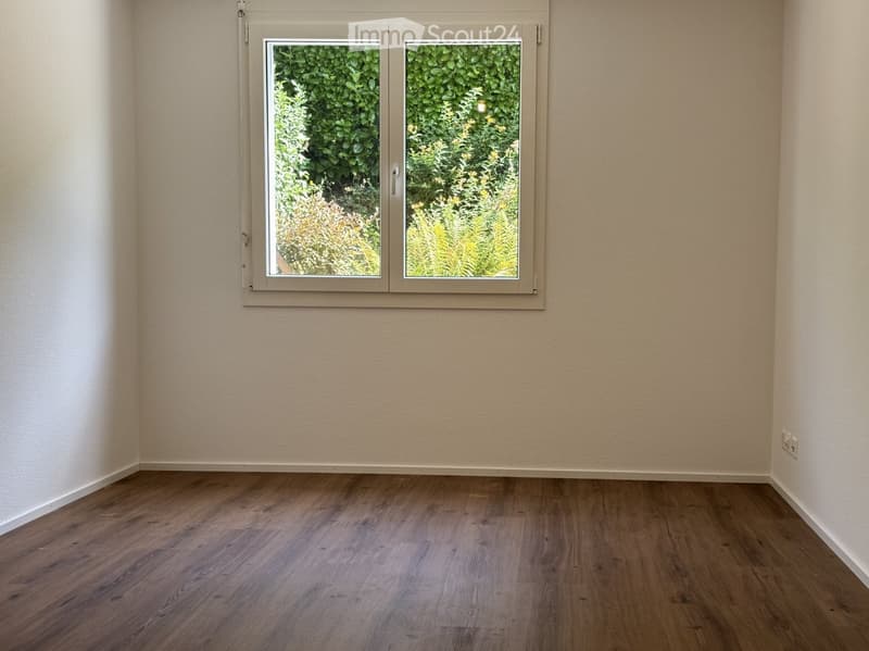 Empty room with light-colored walls and wooden flooring, featuring a large window with a view of greenery outside.