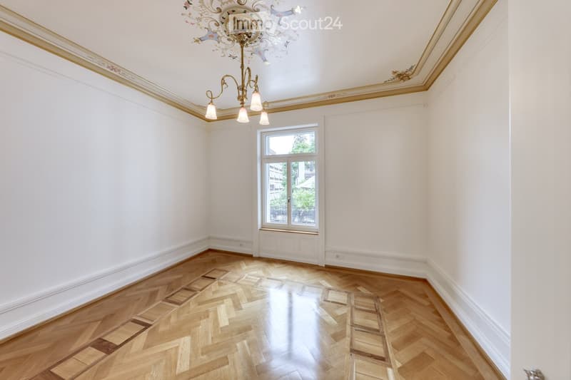 Empty room with white walls, wooden herringbone floor, large window, and ornate chandelier.
