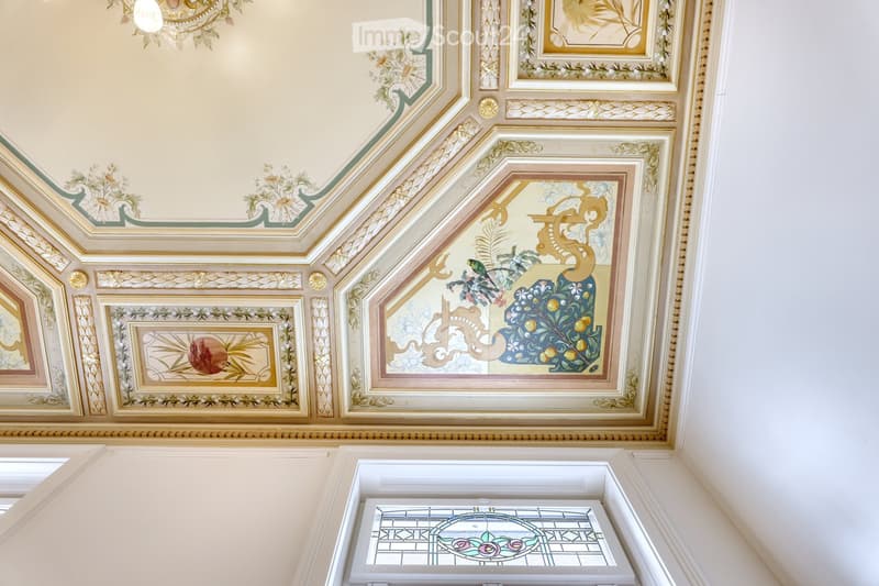 Ornate ceiling with intricate painted designs and gold detailing, featuring floral and geometric patterns, with a stained-glass window below.