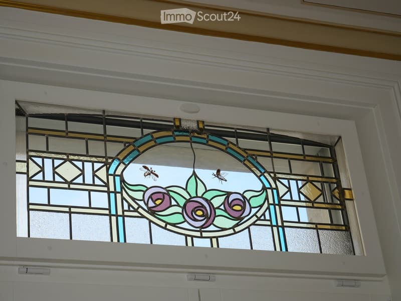 A decorative stained-glass window featuring floral designs and insects, set within a white-framed doorway with a ceiling featuring wooden trim and recessed lighting.