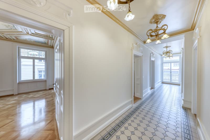 Elegant hallway with polished wooden flooring, decorative ceiling moldings, ornate chandeliers, and stained-glass windows, leading to a tiled corridor with intricate patterns.