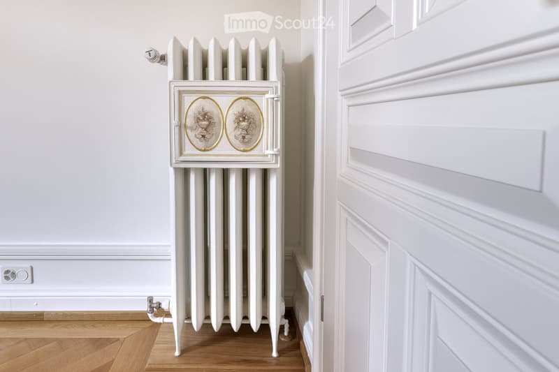 A narrow hallway with white walls, wooden flooring, and a decorative white radiator with ornate gold accents on the left side.