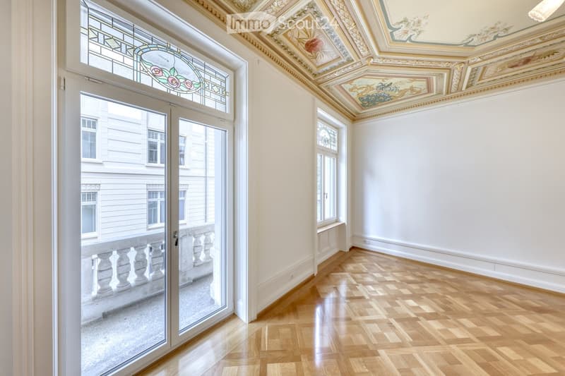Empty room with wooden parquet flooring, white walls, and ornate ceiling. Large windows and a glass door lead to a balcony area.