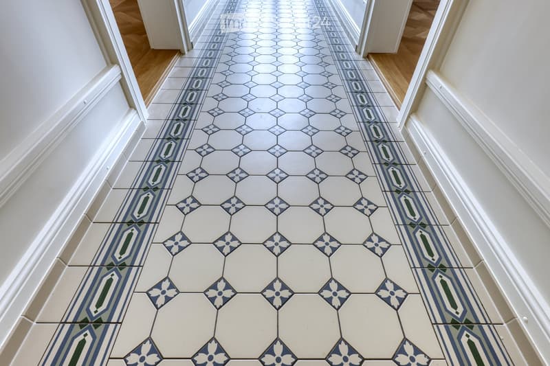 Hallway with hexagonal tile floor in white and blue, featuring floral and geometric patterns, white walls, and wooden paneling at the ends.