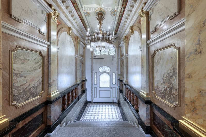 Ornate hallway with marble walls, decorative moldings, chandeliers, and a patterned tile floor leading to a white door.
