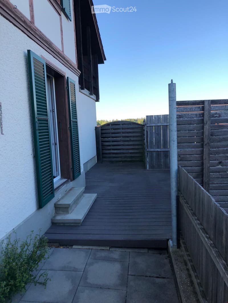 A wooden pathway leads to a gated entrance, flanked by a white building with green shutters and a wooden fence on the right.