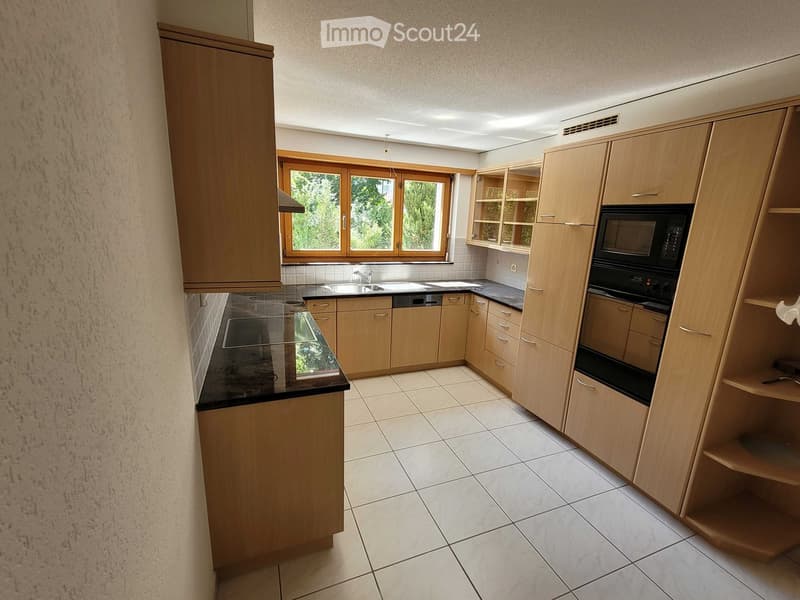 Modern kitchen with light wooden cabinets, black countertops, and stainless steel appliances including a microwave, oven, and dishwasher. The space features a large window with wooden frames and tiled flooring.