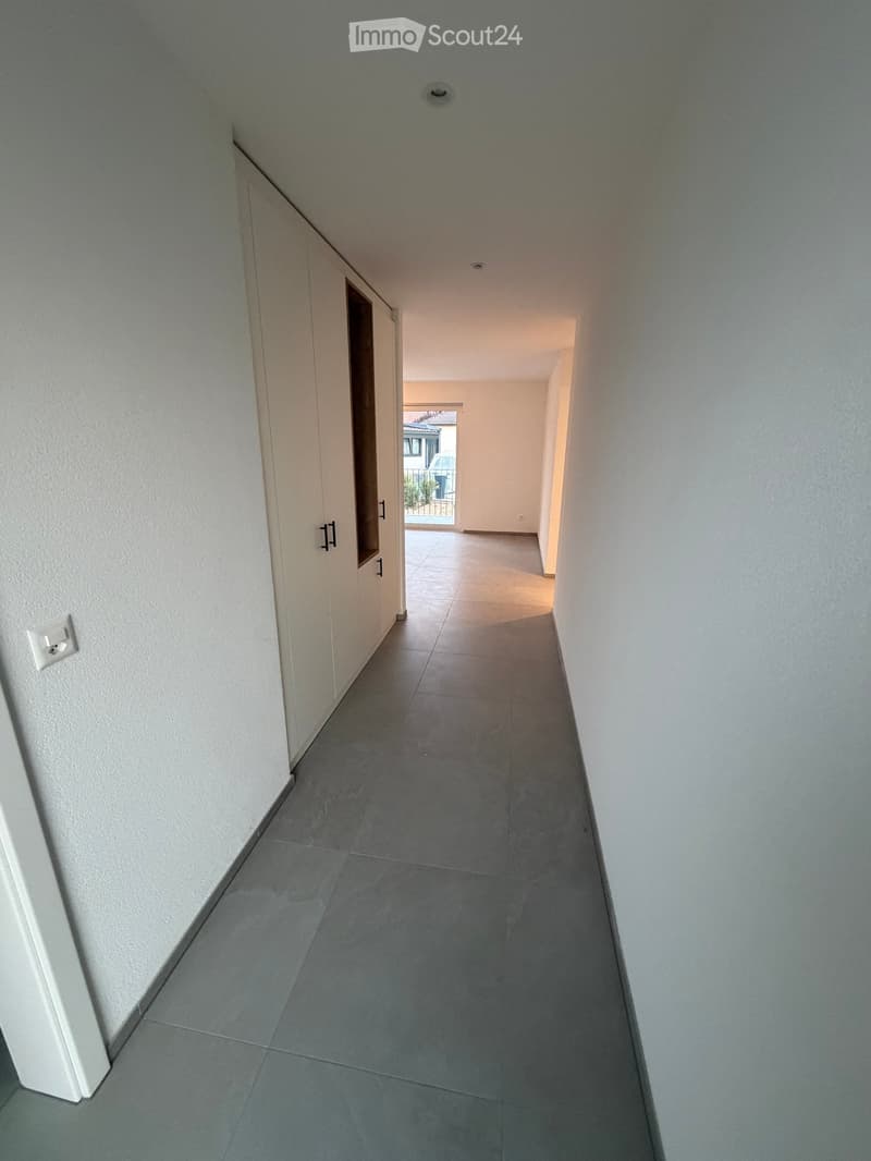 Clean, modern hallway with grey tiled flooring and white walls, featuring a closed white cabinet on the left and an open doorway leading to another room.