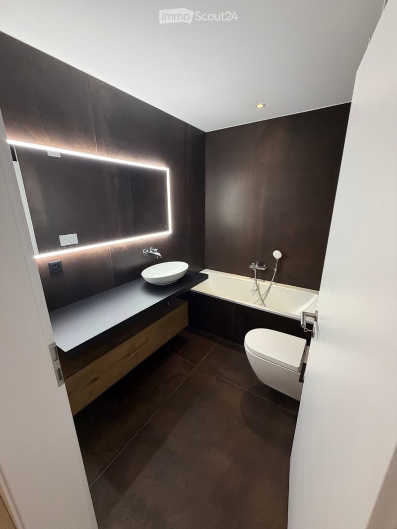 Modern bathroom with dark wooden walls, a white sink, bathtub, toilet, and a large illuminated mirror above the sink.