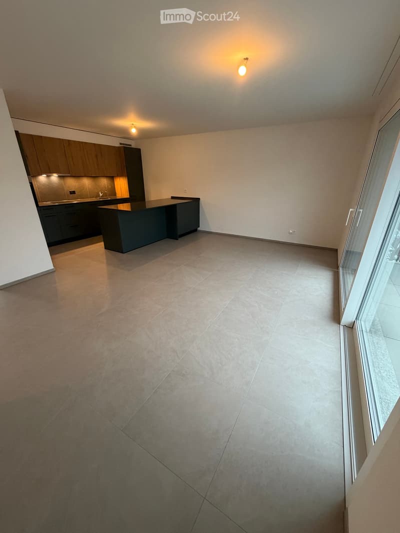 Empty room with grey tiled floor, white walls, and a dark kitchen area with cabinets and a countertop visible in the background.