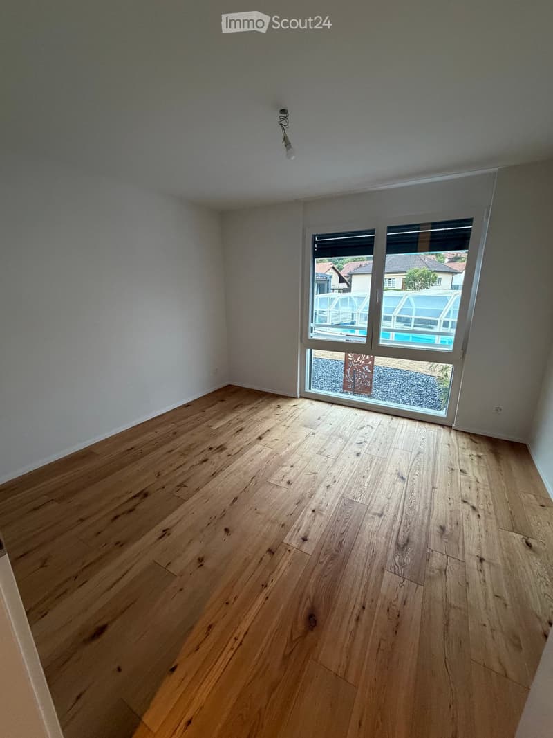 Empty room with wooden flooring and large windows overlooking a pool and garden area.