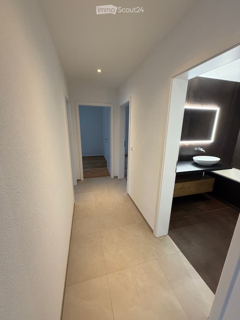 Modern hallway with beige tiled flooring, white walls, and recessed lighting. Features a partially visible bathroom with a dark vanity, sink, and illuminated mirror on the right side.