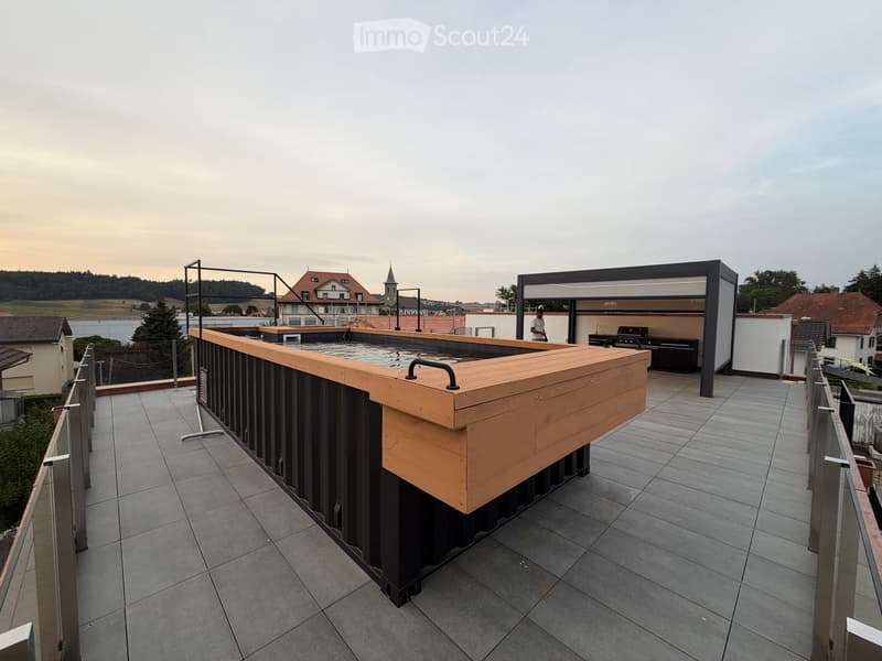 Modern rooftop terrace with a wooden bar, built-in grill, and a covered seating area, overlooking a scenic town and river view.