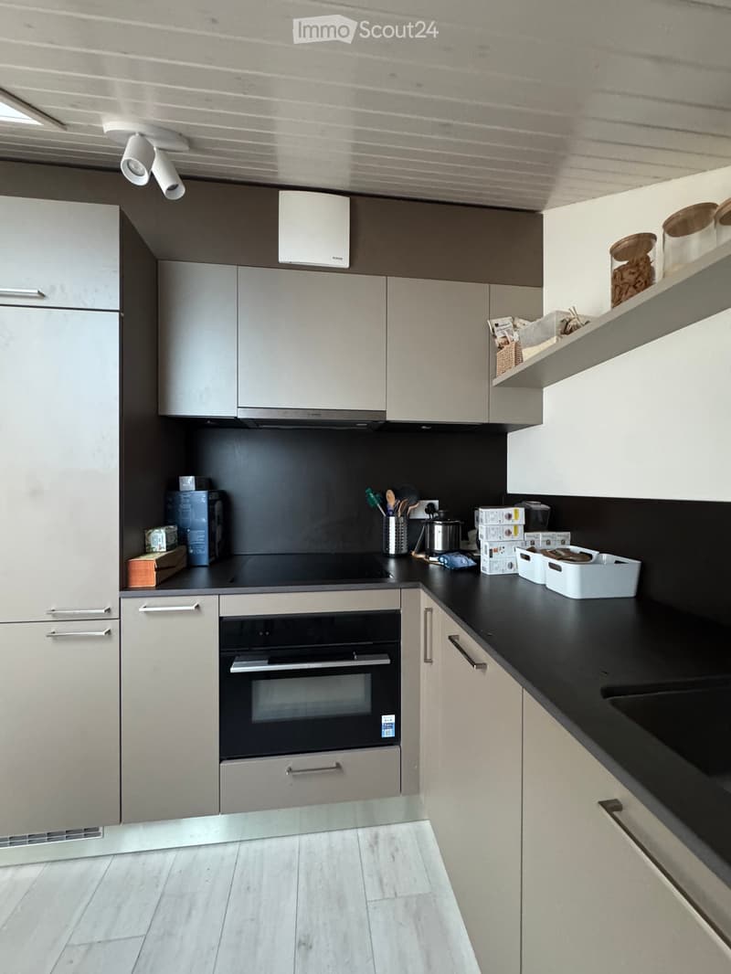 Modern kitchen with light grey cabinets, black countertops, and stainless steel appliances including a refrigerator, oven, and sink. Shelves hold jars and kitchen items.