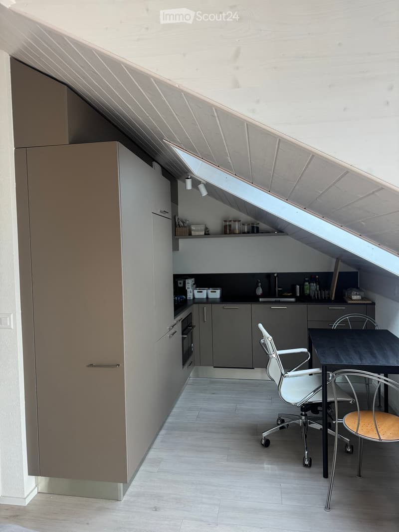 Modern kitchen with grey cabinets, white walls, and a slanted wooden ceiling. Features a black countertop, sink, and a small dining area with a table and chairs.