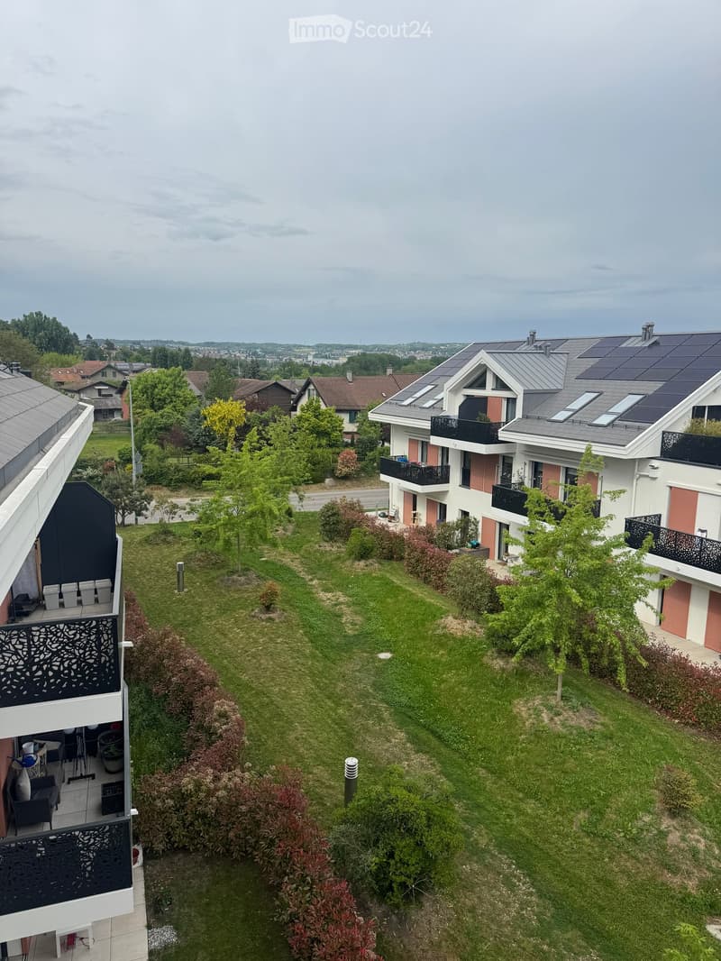 A well-maintained garden with a lawn, shrubs, and trees, surrounded by modern residential buildings with balconies and solar panels on the roofs.