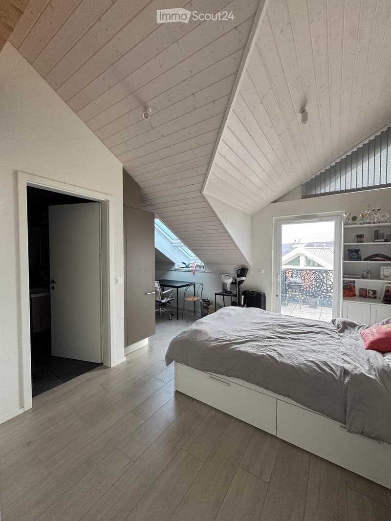 Modern attic bedroom with wooden slanted ceilings, large windows, and built-in storage. Features a bed, chair, table, and shelves.