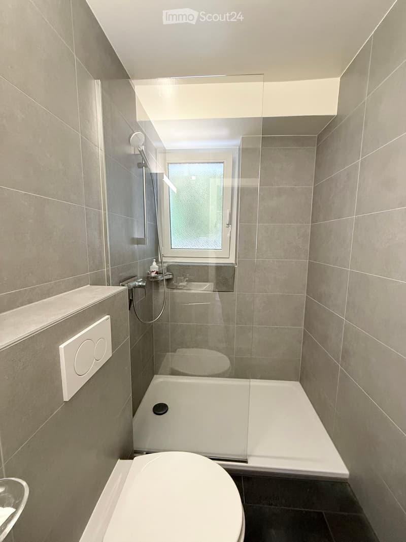 Modern bathroom with a bathtub, toilet, shower, and a frosted window. The walls are tiled in a neutral gray color, and the floor is dark gray.