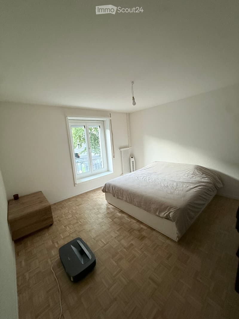A well-lit bedroom with a wooden floor, a bed with a grey duvet, a nightstand, a radiator, and a vacuum cleaner on the floor.