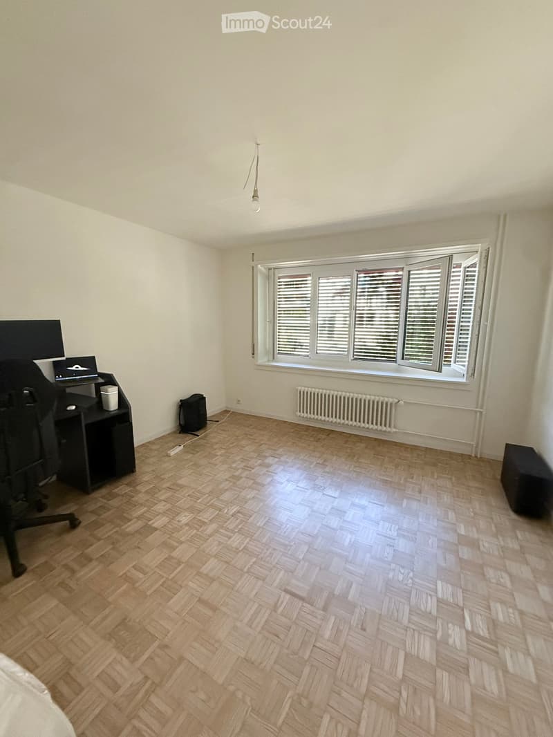 A modern, unfurnished room with wooden herringbone flooring, white walls, and large windows with blinds. A desk with a laptop and chair is positioned near the window, and a radiator is mounted below the window. A small black stool is placed near the right