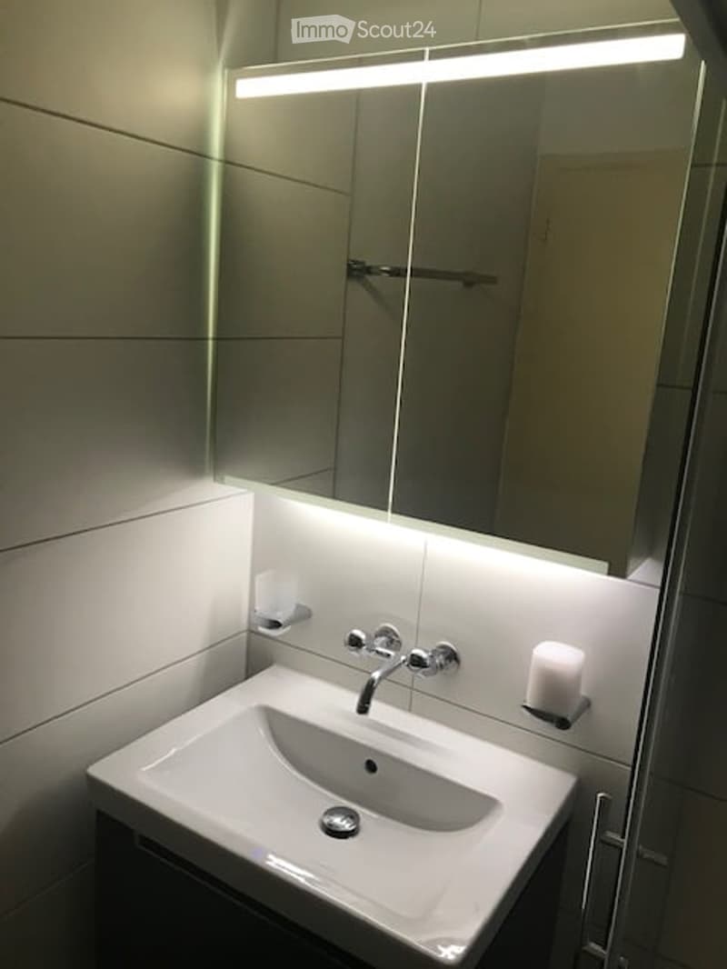 Modern bathroom with a white sink, sleek faucet, large mirror, integrated lighting, and a decorative candle on the counter.