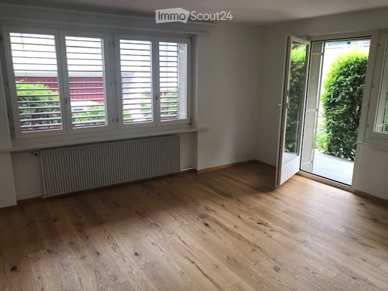 Empty room with wooden flooring, large windows with shutters, radiator, and an open door leading to a balcony or outdoor area.
