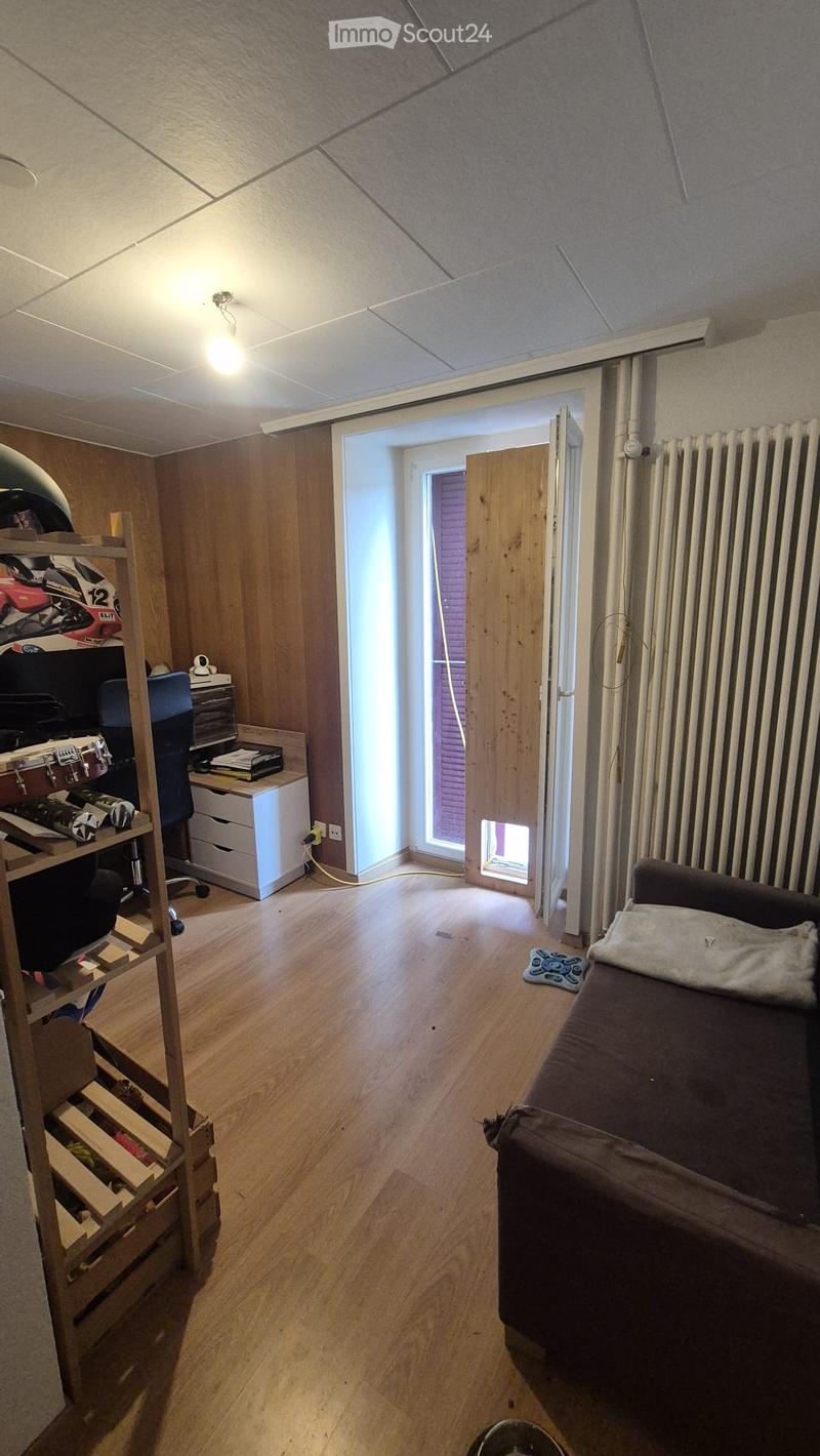 A furnished room with wooden walls and flooring, featuring a sofa, a desk with a chair, a shelf with helmets and a drum, and a small opening leading to another space.