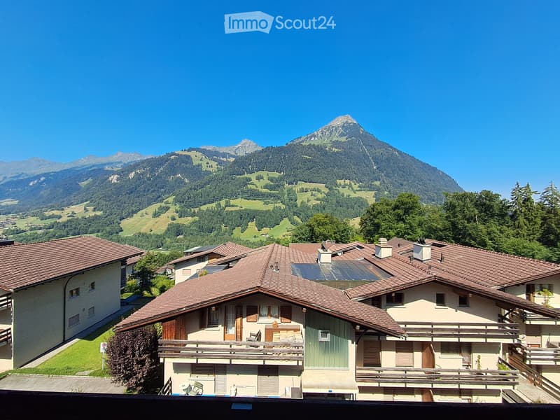 A cluster of houses with red-tiled roofs and balconies, surrounded by lush greenery and a scenic mountain backdrop.