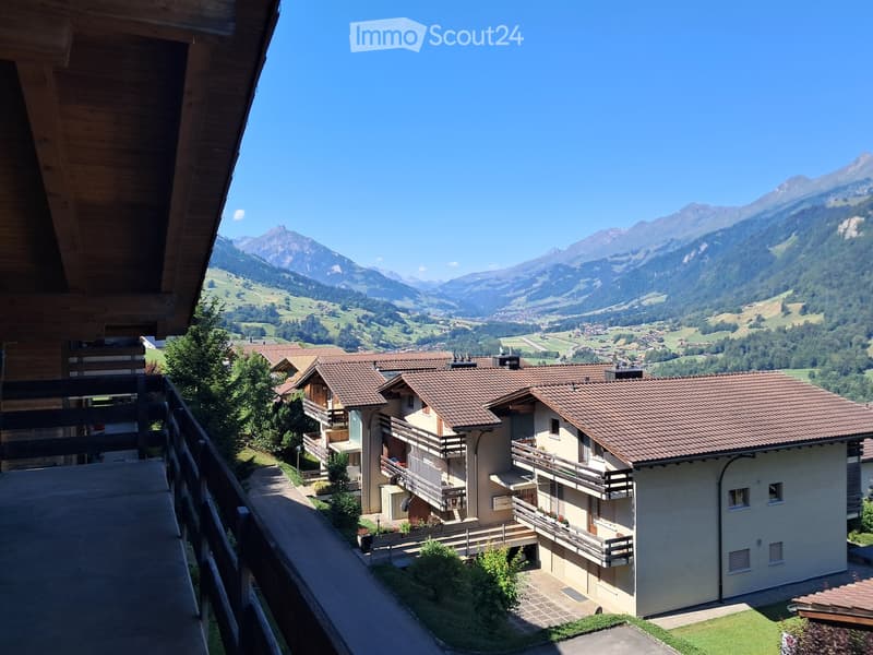 A multi-story residential building with balconies and a tiled roof, situated in a picturesque valley with mountain views.