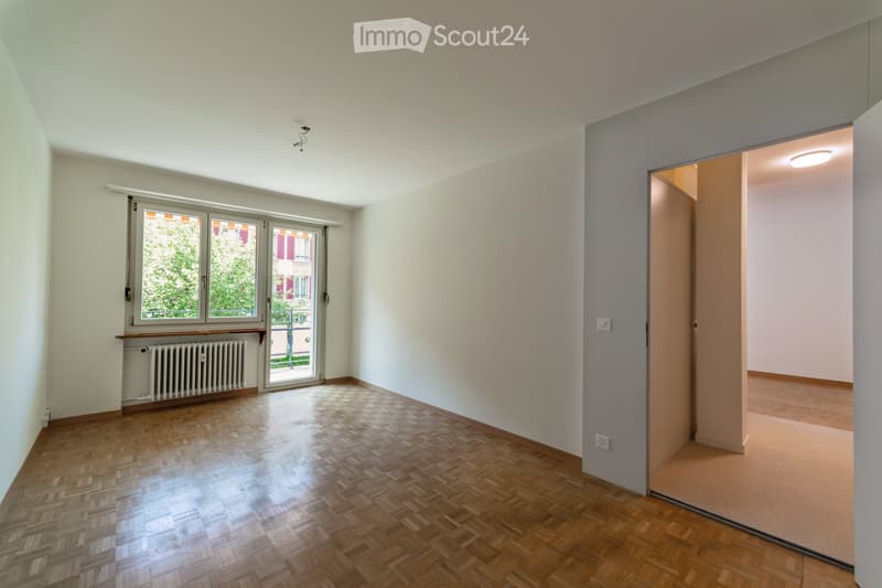 Empty room with wooden flooring, white walls, large window with blinds, radiator, and an open doorway leading to another room.