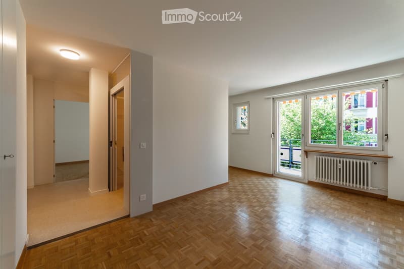Empty room with wooden herringbone flooring, white walls, and large windows with blinds. Features a radiator and a door leading to another room.