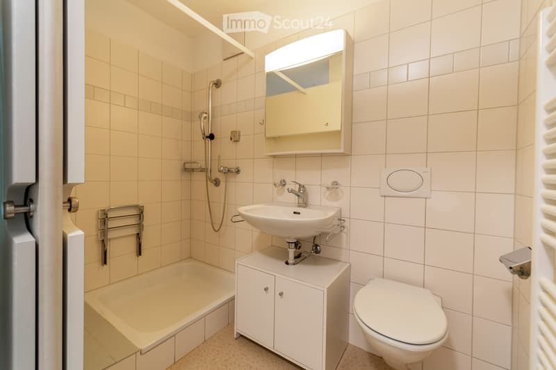 Modern bathroom with white tiles, a bathtub, a sink with a mirror, and a toilet. Features include a handheld showerhead, a towel rack, and a built-in cabinet under the sink.