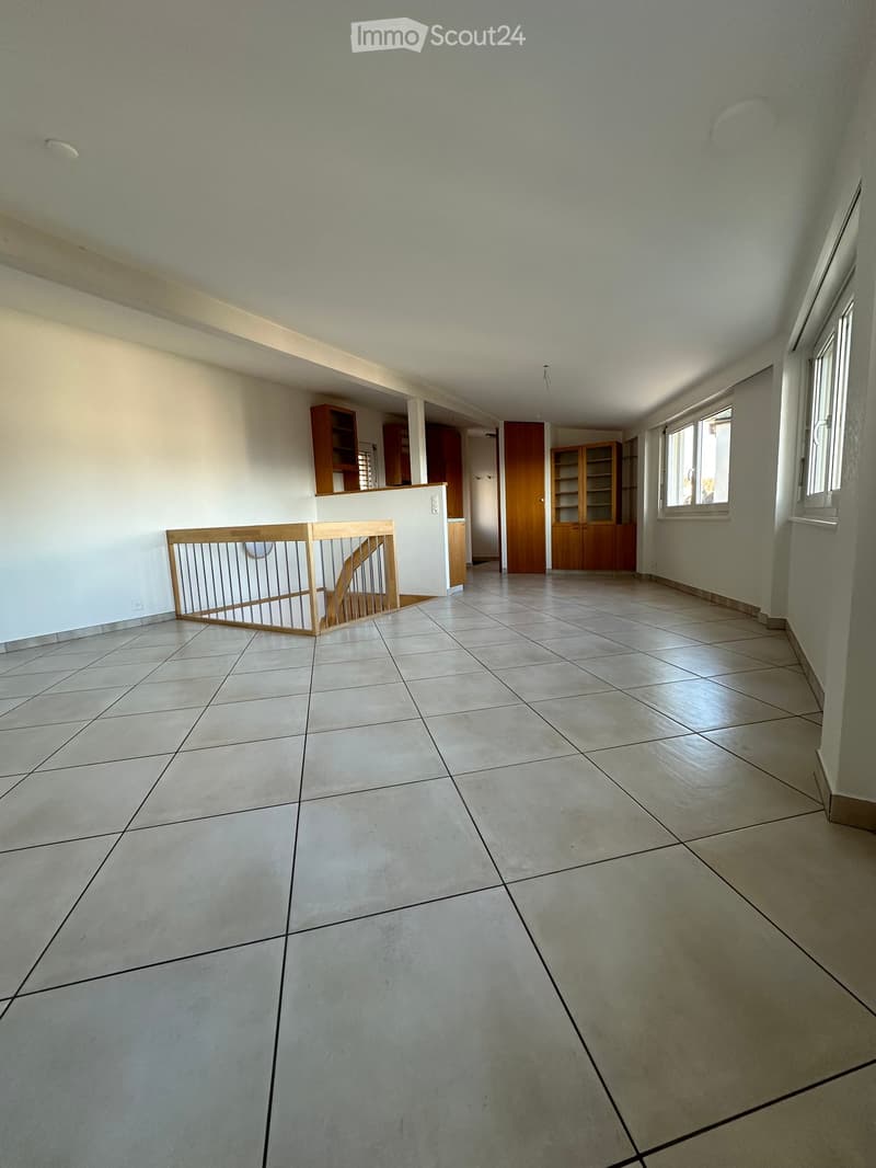 Empty room with tiled floor, white walls, and wooden furniture visible in the background, including cabinets and a staircase railing.