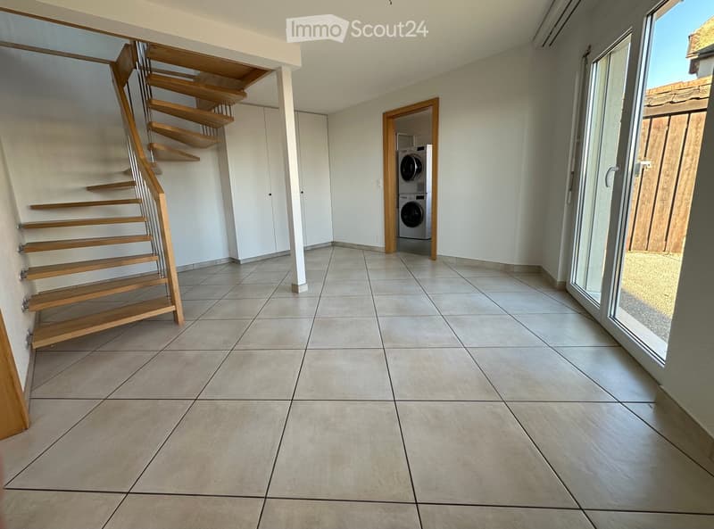 Empty room with tiled floor, wooden staircase, white walls, and a washing machine in the corner.