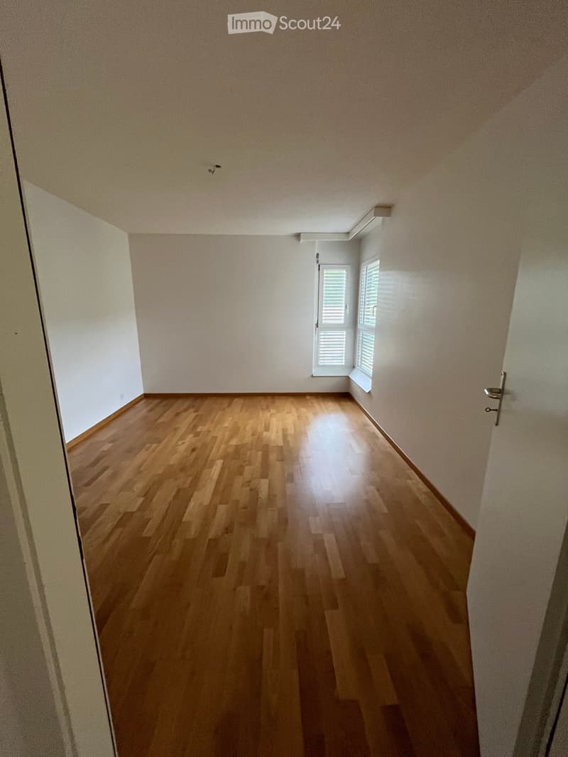 Empty hallway with wooden flooring and white walls, featuring a window with blinds at the end.