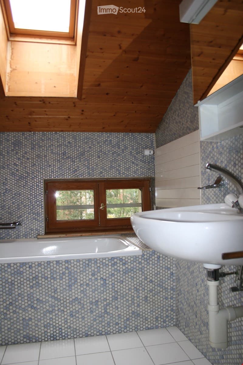 Bathroom with a white bathtub, a white sink, and a chrome faucet. The walls are covered with blue and white mosaic tiles, and the ceiling is wooden with skylights. There is a window with wooden frames and a view of trees outside.