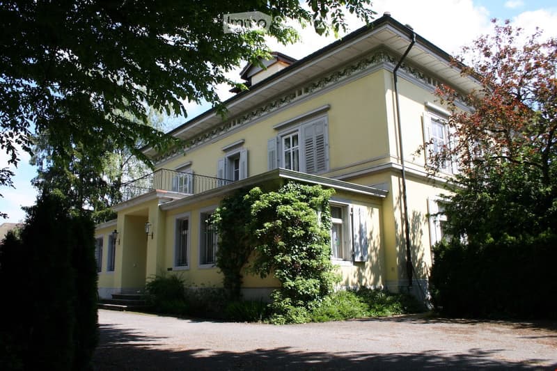 Two-story yellow house with white shutters, balcony, and surrounding greenery.