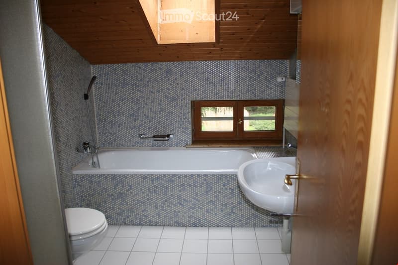 Bathroom with a bathtub, sink, toilet, and a shower hose. The walls are tiled with a blue mosaic pattern, and the ceiling is wooden with skylights. There is a window with wooden frames offering a view of greenery outside.