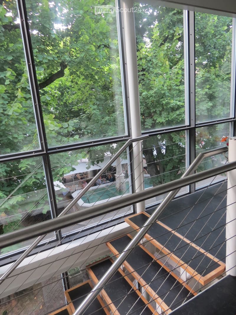Modern hallway with large glass windows overlooking a green outdoor area, featuring a sleek metal railing and wooden steps.