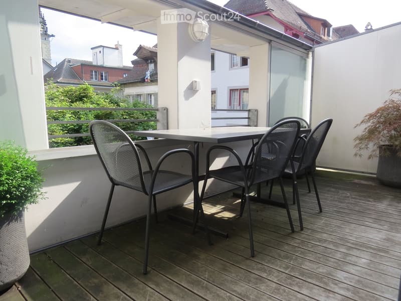 Balcony with wooden decking, metal table and chairs, potted plants, and a view of neighboring buildings.