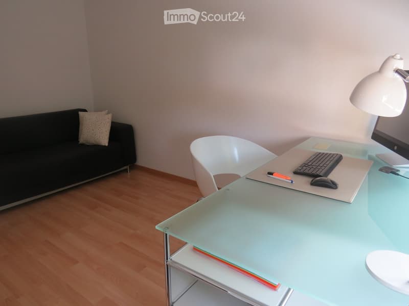 Modern furnished room with a black sofa, white chair, glass desk, computer setup, and a floor lamp.