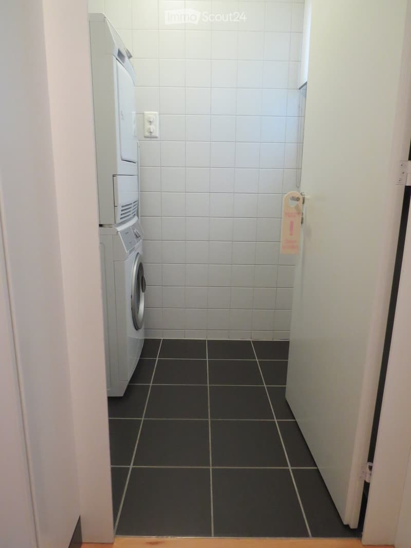 A narrow hallway with white tiled walls and black tiled floor, featuring a washing machine on the left side and a door with a sign on the right.