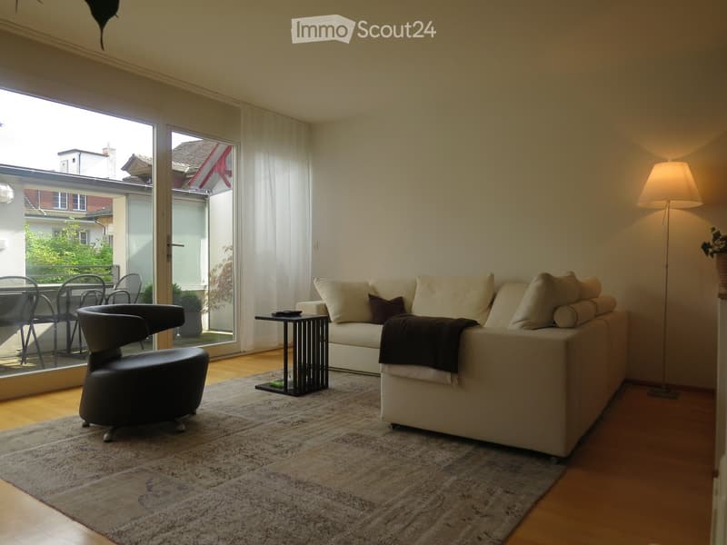 Bright living room with large windows and a balcony, featuring a cream-colored sofa, a black leather chair, a small black side table, a floor lamp, and a patterned rug.