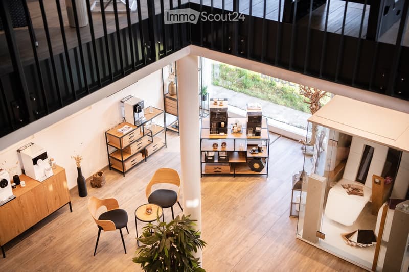 A modern, open-plan space with wooden flooring, featuring a seating area with chairs and a small table, a coffee station with a coffee machine and cups, and a small cabinet with drawers and a vase.